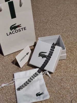 Silverfärgat armband från Lacoste - Oanvänd(nyskick )Stilrent armband i silverfärgad metall från Lacoste med klassisk krokodil-logga. Armbandet har en länkdesign och levereras med originalförpackning, tygpåse och kort. Perfekt accessoar för att lyfta din outfit.ett till ett, bra present , fraktar snabbt