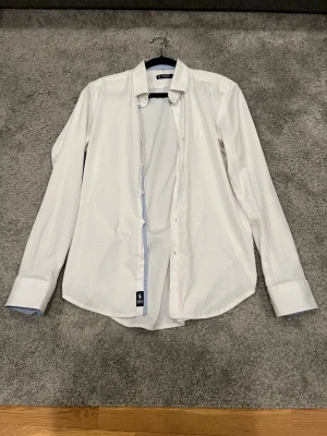 Ralph Lauren skjorta  - Klassisk vit skjorta från Polo Ralph Lauren med diskret logga broderad på bröstet. Skjortan har lång ärm, knappar framtill och är tillverkad i bomull. Perfekt till både jeans och chinos för en clean look.