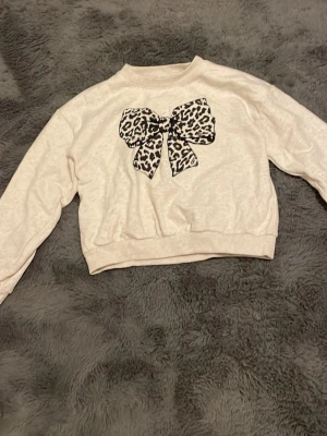  Beige tröja med leopardrossett - Supersöt vit sweatshirt med en stor rosett framtill i leopardmönster. Tröjan har croppad passform och långa ärmar. Perfekt för dig som gillar statement-detaljer och djurmönster.