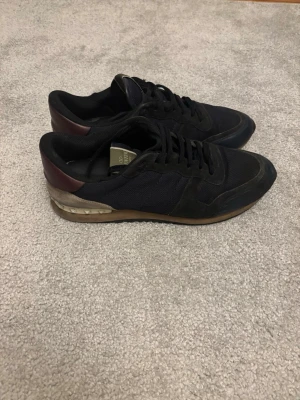 Valentino Rockrunner svarta sneakers - Snygga svarta Valentino Rockrunner sneakers med detaljer i vinrött och olivgrönt. Ovandelen är i mesh och mocka, med klassisk snörning och platt sula. Bakpartiet har en cool metallisk detalj och kontrastfärg. Perfekta för dig som vill ha en stilren men unik sneaker.