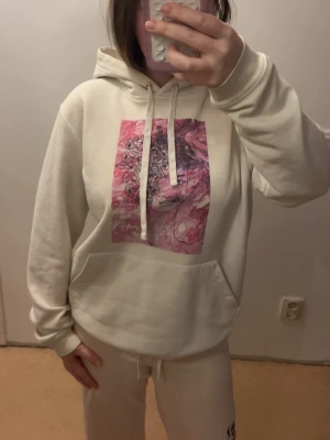 Vit hoodie med rosa tryck - Säljer en vit hoodie med huva och dragsnören. På bröstet finns ett stort grafiskt tryck i rosa och lila toner. Hoodien har en klassisk känguruficka framtill och ribbade muddar vid ärmar och midja. Perfekt för en chill och avslappnad stil.