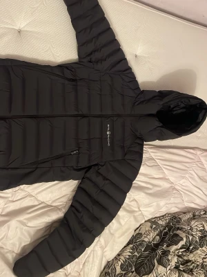 Svart dunjacka från sail racing - Snygg svart dunjacka från sail racing med huva och dragkedja framtill. Jackan har en quiltad design och är perfekt för kalla dagar. Klassisk passform och diskret logga på bröstet. Perfekt för dig som vill hålla värmen med stil.