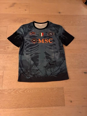 Svart camo t-shirt med tryck MSC - Cool svart och mörkgrå t-shirt med camouflagemönster och tryck i orange och vitt. Framtill står det MSC och flera patch-liknande tryck, baktill finns ett spindelnät och texten SO.GESANA. Perfekt för dig som gillar unika och sportiga plagg.