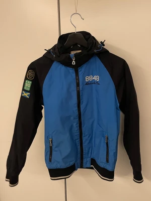 Jacka 8848 Altitude  - Snygg blå och svart vindjacka från 8848 Altitude med huva, dragkedja framtill och två fickor med dragkedja. Jackan har broderade detaljer och patchar på ärmen samt ribbade muddar vid ärmslut och nederkant. Perfekt för blåsiga dagar.