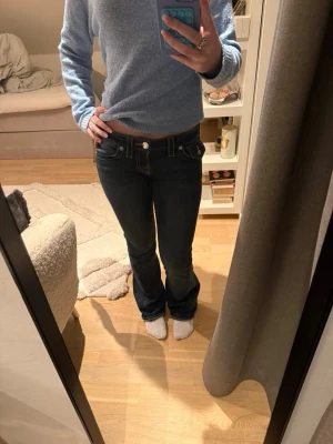 Mörkblå True Religion jeans bootcut - Säljer ett par mörkblå jeans från True Religion med bootcut-modell och låg midja. Jeansen har tydliga kontrastsömmar, snygga fickdetaljer med knappar bak och klassisk logopatch. Tillverkade i jeansmaterial med en cool, lite faded look.
