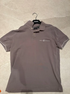 Grå pikétröja från Sail Racing - Snygg grå pikétröja från Sail Racing med klassisk krage och knappar framtill. Tröjan har diskret logga broderad på bröstet och vita detaljer vid ärmsluten. Perfekt för en stilren och sportig look.