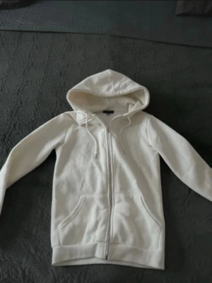 Vit zip-hoodie med fickor - Snygg vit hoodie med dragkedja. Klassisk modell med huva och dragsko samt två stora fickor framtill. Perfekt för en chill och avslappnad stil. Mjuk bomullskänsla och ribbade muddar vid ärmslut och nederkant.