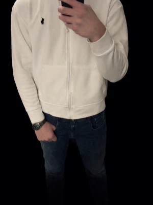 Vit hoodie från Ralph Lauren - Snygg vit hoodie från Ralph Lauren med dragkedja och klassisk logga på bröstet. Hoodien har fickor framtill och ribbade muddar vid ärmslut och nederkant. Perfekt för en clean och stilren look.