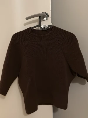 Mörkbrun stickad kortärmad tröja från ZARA - Snygg mörkbrun stickad tröja med rund hals och trekvartsärmar. Tröjan har ett diskret ribbstickat mönster och är gjord i ett mjukt material som känns skönt mot huden. Perfekt för lager på lager och enkel att matcha med olika outfits.