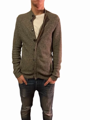 Grå kofta från Massimo Dutti, M - Snygg gråmelerad kofta från Massimo Dutti i bomull. Modellen har lång ärm, ribbade muddar och knappar framtill. Perfekt för dig som gillar stilrena och klassiska plagg med lite struktur. Passar dig som vill ha en normal passform och gillar neutrala färger.