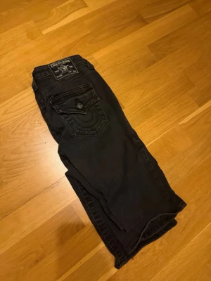 Svarta jeans från True Religion - Svarta jeans från True Religion med klassisk bakficka och tydliga sömmar. Jeansen har en snygg passform och är tillverkade i jeansmaterial. Perfekta för dig som vill ha en stilren look med lite edge.