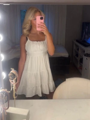 Vit somrig klänning med volanger - Söt vit kortklänning med tunna axelband och volangdetaljer. Klänningen har en luftig passform och är perfekt för varma dagar. Tillverkad i bomull med diskret broderat mönster för en extra touch.