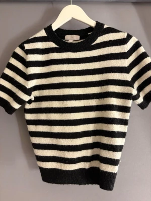 Randig svartvit tröja från H&M - Kortärmad stickad tröja från H&M i svart och vitt med klassiskt horisontellt randigt mönster. Tröjan har rund halsringning och ribbade kanter vid ärmslut och nederkant. Perfekt för en stilren och trendig look.