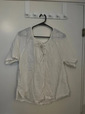 Vit blus med knytning från H&M - Luftig vit blus från H&M med korta ärmar och dekorativ knytning vid halsen. Blusen har en avslappnad passform och är tillverkad i ett lätt tyg som passar perfekt till sommaren. Enkel att matcha med både jeans och kjol.