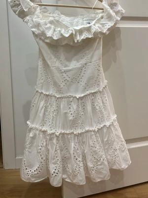 Vit offshoulder klänning med volang från Zara  - Superfin vit kortklänning med offshoulder och volangdetaljer. Klänningen har broderade spetsmönster och flera lager med rynkade volanger. Använd ungefär 2 ggr. Perfekt för sommarens alla tillfällen och till studenten! 