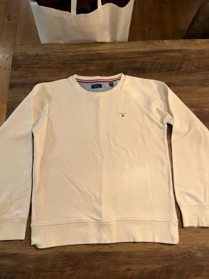 Vit sweatshirt från GANT - Säljer en stilren vit sweatshirt från GANT med broderad logga och sköld på bröstet. Tröjan har rund halsringning med ribbad kant och en diskret röd och marinblå rand på insidan av kragen. Tillverkad i mjukt bomullsmaterial och har långa ärmar.