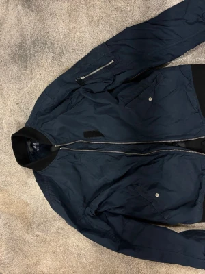 Mörkblå bomberjacka från H&M - Snygg mörkblå bomberjacka från H&M med klassisk ribbad krage, dragkedja framtill och praktiska fickor med tryckknappar. Jackan har en extra ficka med dragkedja på ärmen och ribbade muddar vid ärmslut och nederkant. Perfekt för en cool och avslappnad stil.