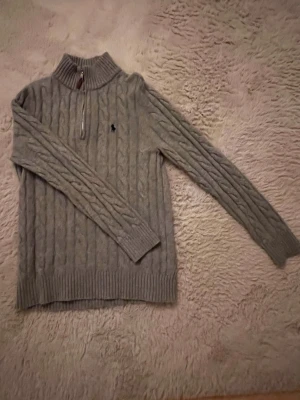 Grå kabelstickad tröja Polo Ralph Lauren - Kabelstickad grå tröja från Polo Ralph Lauren med halv dragkedja och hög krage. Tröjan har långa ärmar, ribbade muddar och den klassiska broderade loggan på bröstet. Perfekt för dig som gillar stilren och tidlös design.