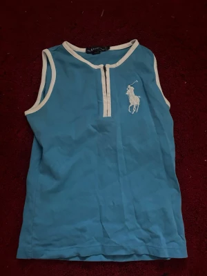 Blå ärmlös topp från Polo Ralph Lauren - Snygg blå ärmlös topp från Polo Ralph Lauren med vit kontrastkant och dragkedja framtill. Stor broderad logga på bröstet. Perfekt till sommaren och enkel att matcha med jeans eller kjol.