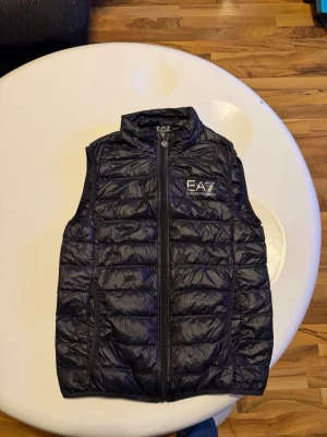 Svart dunväst från EA7 Emporio Armani - Snygg svart dunväst från EA7 Emporio Armani med quiltad design och hög krage. Västen har dragkedja framtill och två sidofickor. Perfekt för lager-på-lager och har en stilren logga på bröstet. Lätt och smidig att bära till olika outfits. Lappen bortklippt köpt från zalando använd fåtal gånger fint skick! Passar er med storlek xs-s. Bara skriva vid frågor!