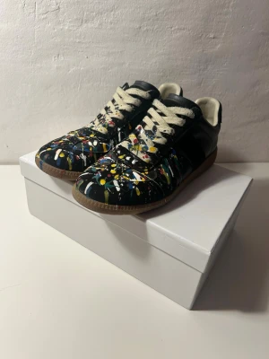 Maison Margiela sneakers svart färgstänk - Unika sneakers från Maison Margiela i svart skinn med färgstänk i vitt, gult, rött och grönt. Klassisk låg modell med vita snören och brun gummisula. Perfekta för dig som vill sticka ut med en kreativ och trendig look.