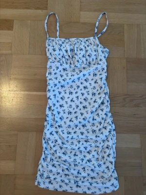 Vit blommig kortklänning med smala band - Söt vit kortklänning med blått blommönster och smala axelband. Klänningen har rynkade detaljer och en figurnära passform. Perfekt för varma dagar och sommarens alla tillfällen. Storlek "Teen" motsvarande 32.