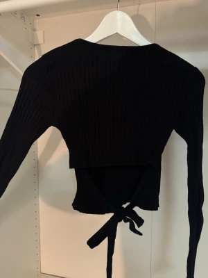 Svart ribbad omlottopp från FB Sister - Svart långärmad topp från FB Sister / New yorker i ribbad bomullsmix. Omlottmodell med knyt i ryggen som ger en snygg, tajt och trendig look. Perfekt att styla med jeans eller kjol för en cool outfit.😍