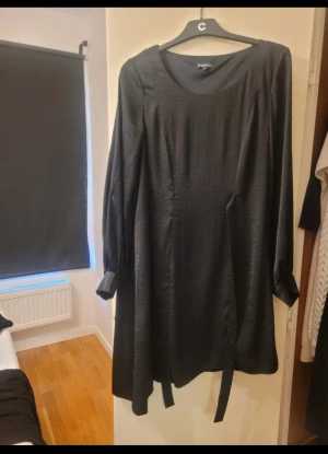 Svart långklänning från Stockh LM - Svart långklänning från Stockh LM i storlek 36. Klänningen har rund halsringning, långa ärmar med knyt vid ärmslut och ett bälte i midjan. Materialet har en lätt glansig yta som ger en elegant känsla. .