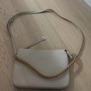 Beige axelväska med flätat handtag +1 - Snygg beige axelväska i slätt skinn med ett flätat handtag i ljusbrun ton. Finns även band som matchar väskan.
