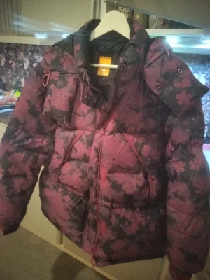 Blommig pufferjacka från Timberland - Säljer en fet pufferjacka från Timberland med blommigt mönster i mörk lila och svart. Jackan har huva, dragkedja och två stora fickor med knappar framtill. Perfekt för dig som vill sticka ut med en unik look i vinter.