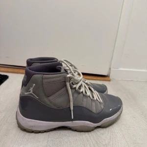 Gråa Nike Air Jordan 11 sneakers - Säljer ett par gråa Nike Air Jordan 11 med vit sula och vita snören. Skorna har en blandning av mocka och glansigt material på ovandelen, samt klassisk Jumpman-logga på sidan. Mellanhöga och riktigt ikoniska för dig som gillar streetwear.