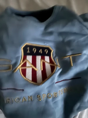 Ljusblå sweatshirt från GANT - Snygg ljusblå sweatshirt från GANT med stort broderat emblem och texten 'American Sportswear' på bröstet. Tröjan har rund hals, ribbade muddar och lång ärm. Perfekt för dig som gillar klassisk och sportig stil.
