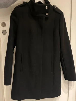 Svart kappa från Lindex med knappar - Säljer en svart kappa från Lindex i storlek S. Jackan har knappar vid axlarna och halsen, samt dold dragkedja. Rak passform, klassisk krage och två fickor framtill. Perfekt för dig som gillar stilrena och tidlösa plagg. Endast använd ett fåtal gånger, fint skick!