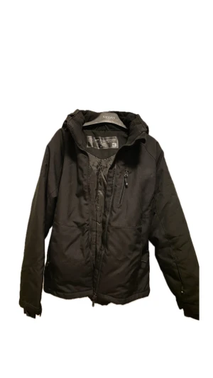 Svart vindjacka från Ski Industries XS - Svart vindjacka från Ski Industries med huva och dragkedja framtill. Jackan har en stilren look, praktisk bröstficka med dragkedja och broderad logga på huvan. Materialet är slitstark polyester och jackan är designad för att vara vind- och vattenavvisande.