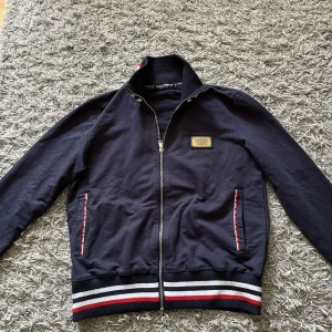 Marinblå kofta från Dolce & Gabbana - Snygg svart jacka från Dolce & Gabbana med dragkedja och ribbade muddar. Jackan har en stilren design med ett märke på bröstet och randiga detaljer i rött och vitt vid ärmslut och nederkant. Säljer för den är för liten för mig, annars för fet