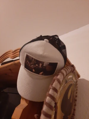 Beige truckerkeps med Maggiore-tryck - Snygg truckerkeps i beige med svart mesh på baksidan. Framtill finns ett stort tygmärke med motiv av en man i kostym och texten 'MAGGIORE'. Kepsen har böjd skärm och justerbar storlek. Perfekt för dig som gillar unika accessoarer med coolt tryck.