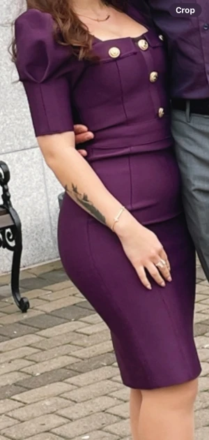 Lila figurnära klänning med guldknappar - Snygg lila klänning med markerade axlar och guldknappar framtill. Klänningen är figurnära med kort längd och har halvlånga ärmar. Perfekt för dig som vill sticka ut med en elegant och modern look.