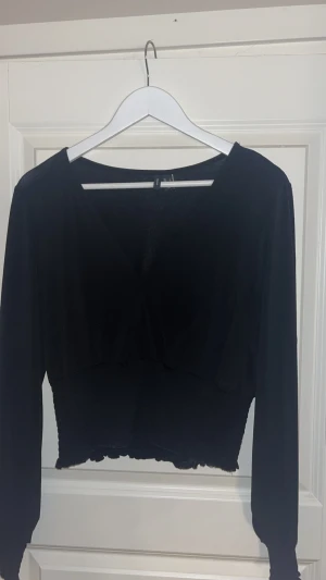 Svart croppad långärmad topp - Säljer en svart croppad topp med lång ärm och v-ringad hals. Toppen har en ribbad och elastisk midja med vågig kant nedtill som ger en snygg detalj. Perfekt att styla med höga jeans eller kjol för en trendig look.