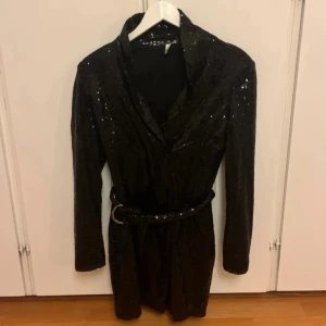 Svart paljettklänning från NLY Trend - Glittrig svart kortklänning från NLY Trend med långa ärmar och bälte i midjan. Klänningen har en snygg krage och är täckt av paljetter som ger en riktigt festlig look. Perfekt för dig som vill sticka ut och glänsa.