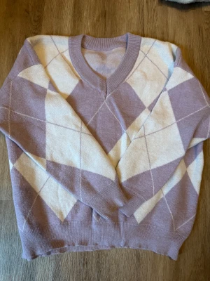 Ljuslila och bege rutig stickad tröja - Säljer en mysig stickad tröja med v-ringning i ljuslila och beige. Tröjan har ett klassiskt rutmönster med ribbade muddar vid hals, ärmslut och nederkant. Perfekt för dig som gillar preppy vibes och vill ha något mjukt och snyggt till hösten.