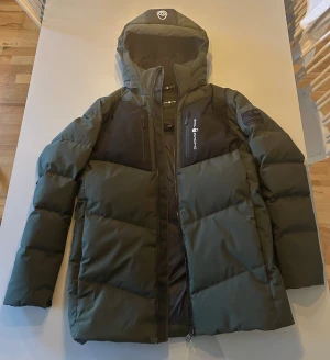 Sail Racing jacka - Säljer en grön dunjacka från Sail Racing i storlek 176, passar XS. Jackan har två svarta bröstfickor med zip. Insidan är fodrad med fjädrar och är perfekt för kalla dagar. Som ny.