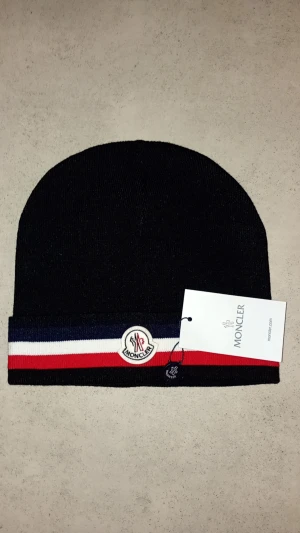 Svart Moncler mössa med ränder - Svart ribbstickad mössa från Moncler med klassiska röda, vita och marinblå ränder vid uppviket. Framtill sitter Monclers broderade logotyp. Mössan är i mjukt material och har en stilren, sportig look.