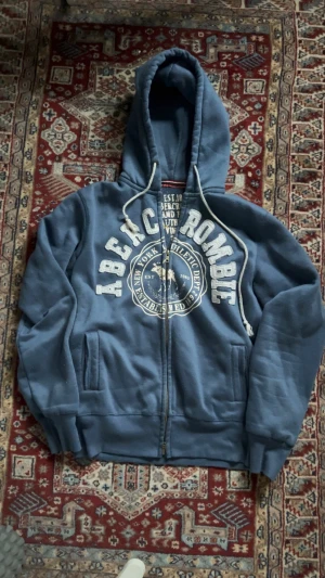 Blå hoodie från Abercrombie & Fitch - Säljer en blå hoodie från Abercrombie & Fitch med stor vit logga och tryck på bröstet. Tröjan har huva med snörning, dragkedja framtill och två fickor. Insidan är mjuk och fleecefodrad, perfekt för kyliga dagar. Klassisk college-vibe och riktigt skön passform.