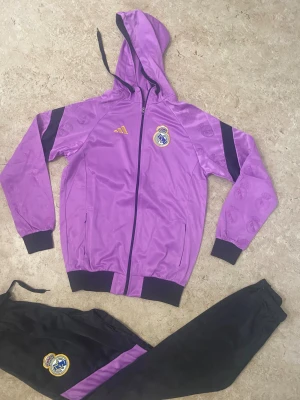 Lila Real Madrid träningsset Adidas - Snyggt Real Madrid träningsset från Adidas med lila huvtröja och svarta byxor. Jackan har dragkedja, huva med snörning och klubbmärke på bröstet. Byxorna har lila detalj och klubbmärke på benet. Perfekt för dig som älskar fotboll och vill sticka ut.