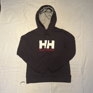 Svart hoodie från Helly Hansen S - Svart hoodie från Helly Hansen i storlek S med stor vit HH-logga och röd text på bröstet. Insidan av huvan är ljusgrå och tröjan har snörning vid huvan. Mjuk och skön bomullsmix, perfekt för chill eller häng. Klassisk passform med långa ärmar och ribbade muddar.