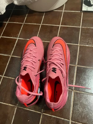 Nike Mercurial Air Zoom rosa fotbollsskor nya - Säljer ett par Nike Mercurial Air Zoom fotbollsskor i en snygg rosa färg med orange och svarta detaljer. Skorna har snörning, platt sula med dobbar och en cool grafisk logga på sidan. Ovandelen är i syntetmaterial för lätt känsla och bra grepp på planen.