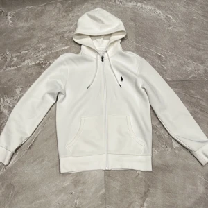 Vit Zip Hoodie Polo Ralph Lauren - Snygg vit hoodie från Polo Ralph Lauren med dragkedja och klassisk logga broderad på bröstet. Tröjan har huva med snörning och två stora fickor framtill. Perfekt för en clean och stilren look.                                           