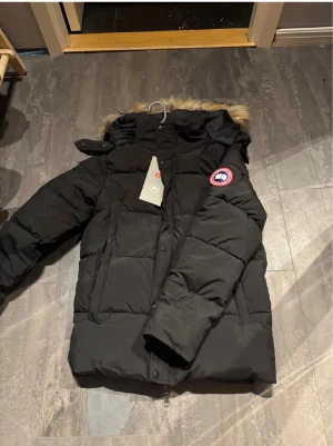 Svart dunjacka från Canada Goose - Säljer en svart dunjacka från Canada Goose med huva och fuskpäls. Jackan har det klassiska Canada Goose-emblemet på ärmen, dragkedja och knappar framtill samt två stora fickor. Perfekt för kalla vinterdagar och riktigt snygg stil . tar även byten