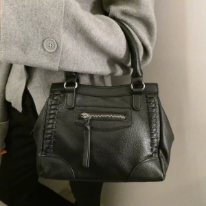 Väska  - Svart väska med detaljer, finns ett litet fack med dragkedja på framsidan, ett lite större på baksidan och 3 fack i väskan.   Två handtag, finns också medföljande justerbart längre axelband