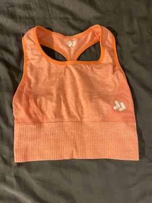Orange sporttopp från 157 - Sportig orange linnetopp från Lager 157 med brottarrygg och ribbade partier nedtill. Tillverkad i stretchigt syntetmaterial som andas, perfekt för träning. Snyggt logotryck framtill och smidig passform som sitter på plats under aktivitet.